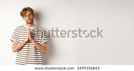 Silly redhead man asking for help, holding hands in pleading gesture and pucker lips, say please while begging you, standing over white background.