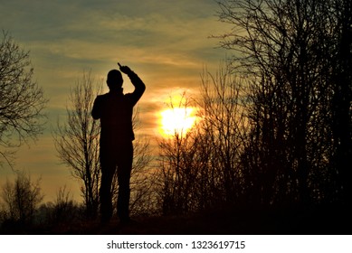 A Silouette Image Of A Man Stood On Top Of Hill Pointing Up At Sky And Sun Amongst Some Trees.