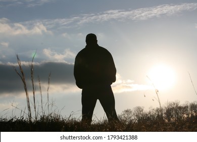 Silouette Of A Man Standing In Front Of A Sun Starting To Set On A Clear Day With Strands Of Grass In The Foreground.