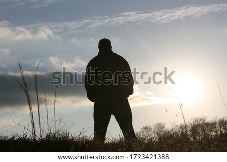 Silouette of a man standing in front of a sun starting to set on a clear day with strands of grass in the foreground.
