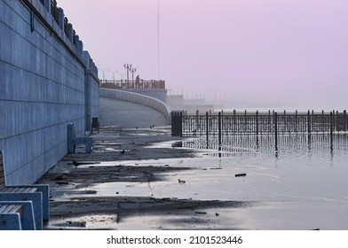 Silt On The Embankment Of The Amur River During A Flood. Heavy Fog At Dawn. Pigeons Walk Through The Puddles. Fencing Lattices. The Figure Of An Athlete In The Background. Blagoveshchensk, Russia.