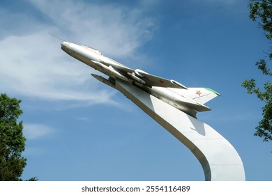 A silver airplane is flying in the sky above a white sculpture. The sculpture is shaped like an airplane wing and is located in a park. The sky is clear and blue, creating a peaceful - Powered by Shutterstock