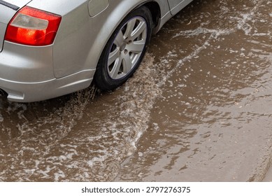 Silver car splashes through muddy puddle. Water swirls around tires, creating foam. Wet pavement reflects car's metallic hue. Scene suggests recent rain or poor drainage. Ideal for weather, urban