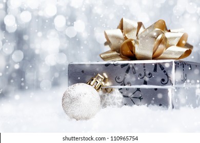 Silver Christmas gift with a huge ornamental golden bow lying with a bauble on a bed of fresh snow with falling snowflakes and copyspace