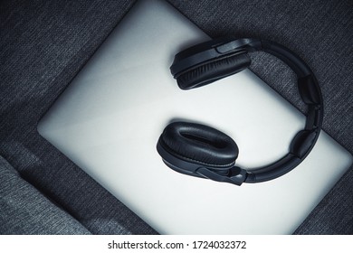 Silver Closed Laptop Computer Sitting On Gray Couch With Big Black Wireless Headphones On Top. 