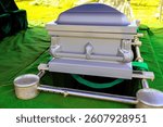 Silver coffin casket is placed above freshly dug grave with green grass, silent surroundings during burial ceremony.