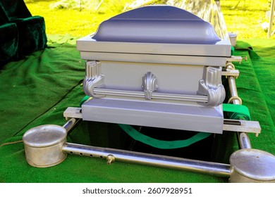 Silver coffin casket is placed above freshly dug grave with green grass, silent surroundings during burial ceremony.