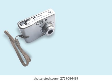 Silver compact digital camera with wrist strap presented in a clean pastel flat‑lay. Close-up view highlights the lens, flash and metallic finish — suitable for technology, travel, product and retro photography themes.
