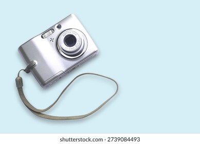 Silver compact digital camera with wrist strap presented in a clean pastel flat‑lay. Close-up view highlights the lens, flash and metallic finish — suitable for technology, travel, product and retro photography themes.