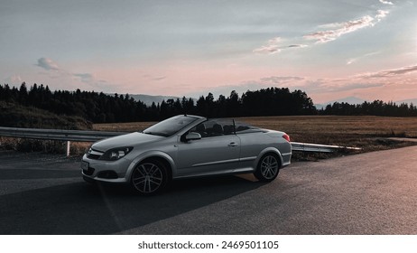 Silver convertible car parked on a paved road during sunset. The background features a scenic landscape with fields, trees, and a colorful sky