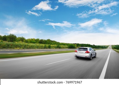 A silver crossover car driving fast on the countryside asphalt road against blue sky with white clouds