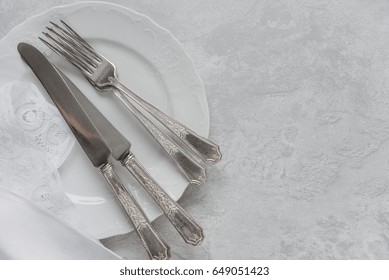 Silver cutlery on a porcelain plate and white napkin with Belgian lace are on the background of gray concrete surface, with copy-space
