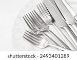 Silver cutlery on a white plate. Close-up of forks and knives set on elegant dishware. Dining background in a restaurant setting. Table arrangement background. Clean shiny metal cutlery.