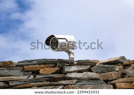 Silver external surveillance camera mounted on stone wall in front of blue sky. Real image.