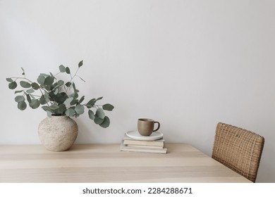 Silver green eucalyptus tree branches in beige textured vase. Brown cup of coffee, tea and old books on wooden table. White wall background. Minimalistic Scandinavian interior, dinning room. Breakfast