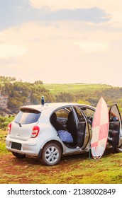 A silver grey car on a cliff with bright pink sirfboard and thermos on roof with its doors open. Surfing lifestyle on tropical island with a small automobile. 