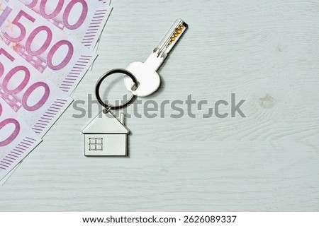 A silver key chain with a house key sits on a white table, with 500 euro bills on the left and plenty of copy space on the right, symbolizing the concept of home purchase and investment.
