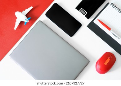 Silver laptop, computer mouse, fountain pen, diary, power bank, mobile phone and toy plane on a red and white table. Concept of preparing for journey, choosing and buying air tickets online. Daylight