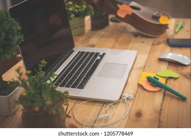 Silver Laptop And Headphone, Cactus Pen, Post Its Notes, Mouse, Mobile Phone And Ukulele On Wooden Working Table With Little Decoration Trees.