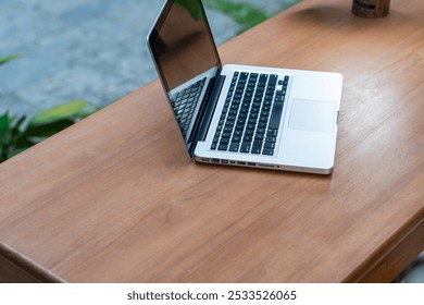 A silver laptop is open on a wooden table, reflecting the surroundings. The background features greenery, suggesting an outdoor or well-lit indoor setting. The laptop's screen is blank - Powered by Shutterstock