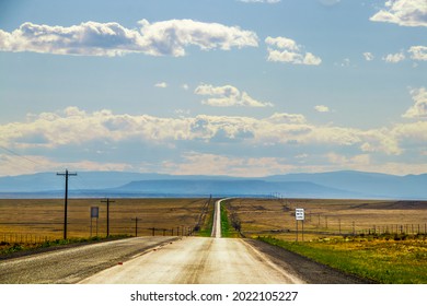 Silver Line Of Two Lane Highway Stretches To Horizon With Blue Layered Mountains In The Distance And Dry Fields  And Electric Lines On Each Side Near Dusk With Pass With Care Sign.