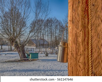 Silver Milk Container  Hanging On A  Column Of A  Wooden Logs Build  Home. In A  Background  A Snow-covered Field With  Willow A  Tree And A Green Doghouse