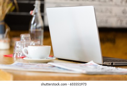 Silver Open Laptop, Cup Of Coffee, Newspaper And Glass Of Water On Wooden Table In Restaurant. Low Angle Perspective, Horizontal Shot.