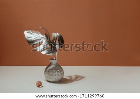 Silver sheet of a monstera in a vase on a background of terracotta wall. Trend art design. Pastel Minimalistic Still Life. Tropical decor concept for modern design