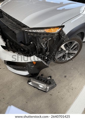 A silver suv with front-end damage and a detached headlight sits in a parking garage after a collision.
