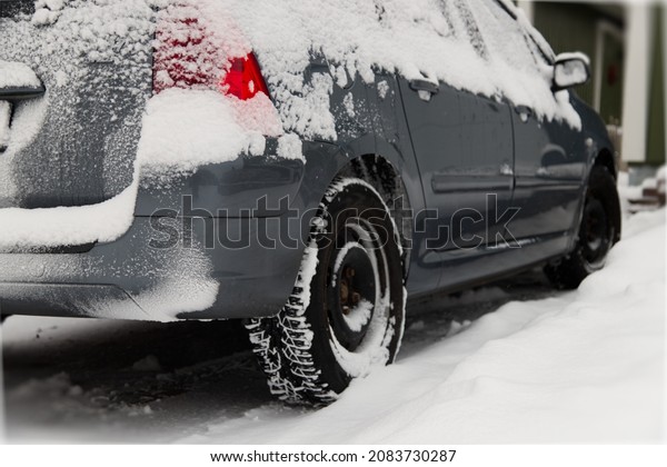 Silver-grey urban car covered with\
snow standing at the side of the road. Image from the backside of\
the car, focus on a back wheel. Snow storm and traffic\
challanges.
