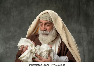 Simeon holding baby Jesus against a dar gray backgroung