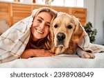 Similarity between dog and owner. Portrait of cheerful woman with blond hair snuggling to adorable golden retriever while lying together on comfy bed under soft blanket.