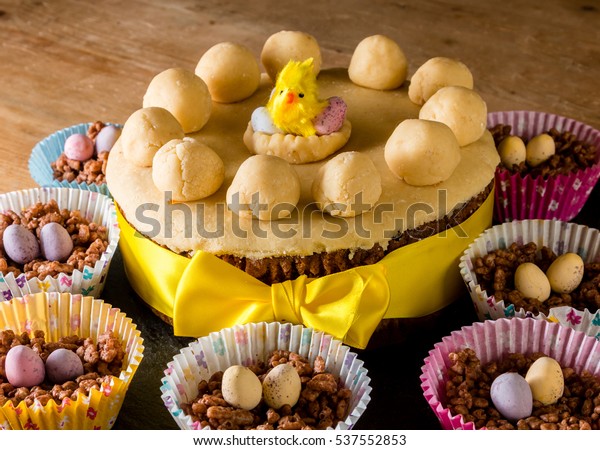 Simnel cake at Easter.  Covered in yellow marzipan\
with a chick toy on top.  The simnel cake has a yellow ribbon and\
is surrounded by chocolate crispy cakes with mini chocolate eggs on\
top