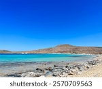 Simos Beach, Elafonissos, Greece. An expansive beach in the Peloponnese with shallow water, white sand and vibrant blue water stretching far out. Sunny blue skies and mountains in the back. 