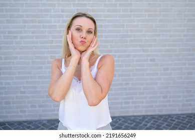 Simpering blond woman holding her hands to her cheeks pursing her lips for the camera - Shutterstock ID 2191656249