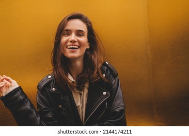 Simpering young brunette woman with a coy smile as she flirts over a gold studio background with copy space. Woman wear black leather jacket and beige shirt is laughing.  - Shutterstock ID 2144174211