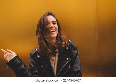 Simpering young brunette woman with a coy smile as she flirts over a gold studio background with copy space. Woman wear black leather jacket and beige shirt is laughing.  - Shutterstock ID 2144546707