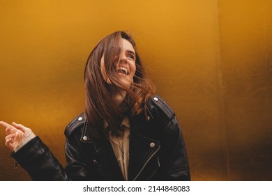 Simpering young brunette woman with a coy smile as she flirts over a gold studio background with copy space. Woman wear black leather jacket and beige shirt is laughing.  - Shutterstock ID 2144848083