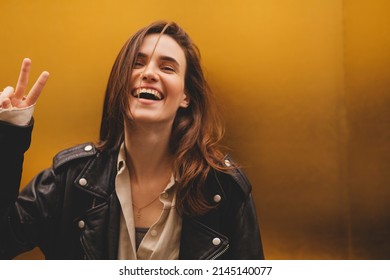 Simpering young brunette woman with a coy smile as she flirts over a gold studio background with copy space. Woman wear black leather jacket and beige shirt is laughing and show peace sign.