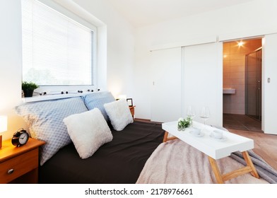 Simple Bedroom In Apartment With Walk-in Closet And Small Bathroom Hidden Behind Sliding Door. In Addition To Bed, There Are Bedside Tables, Lamps, Two Windows And A Tray With Cups On The Bed.