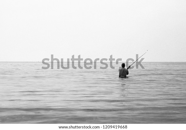 Simple black and white panorama divided in half\
photo of a lonely fisherman on Koh Lanta island, Thailand, fishing\
on the horizon with his spinning line with half of his body\
submerged in water