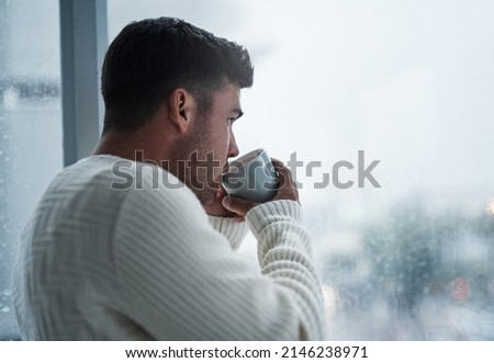 A simple break will bring you back to you. Shot of a young man having coffee and looking out of a window on a rainy day at home.
