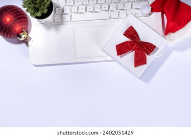 Simple Christmas shopping, online holiday background. White laptop notebook with Santa red hat, gift box, small decorative christmas tree, on white background flatlay top view