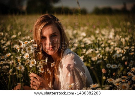 Simple close up portrait of young woman sitting in a daisy meadow wearing white lace playsuit and posing with bunch of daisies.