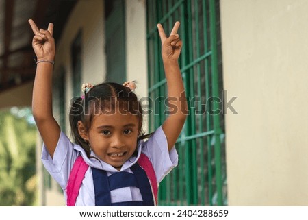 A simple cute preschooler asian girl throws her hands into the air, making peace signs while walking outside her classroom. At a provincial primary school in Bohol.
