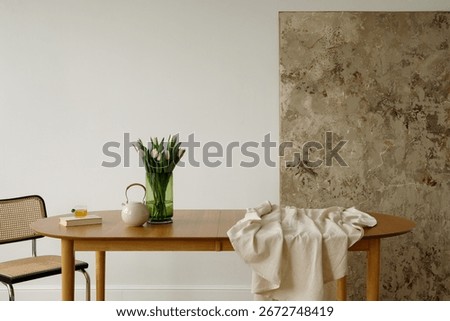 Simple dining table arrangement with white tulips in green vase, ceramic teapot, book, and glass of tea in natural light.