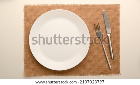 Simple dining table setting with white ceramic plate and silver cutlery over brown placemat on white background. top view, flat lay
