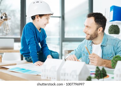 Simple explanation  Pleasant bristled architect explaining the key ideas his project to his son while the boy in hard hat sitting the table   listening to him and smile