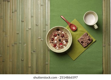 A simple and healthy breakfast arrangement featuring a bowl of granola with dried fruits and white chocolate chips, a granola bar, and a cup of milk on a green placemat with a wooden spoon. - Powered by Shutterstock