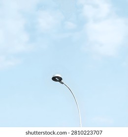 A simple, high-angle minimalist composition showing a sleek, silver curved street lamp post stretching towards a soft pale blue sky filled with light wispy clouds. Perfect for city infrastructure, modern urban fixtures, and minimalist outdoor concepts.